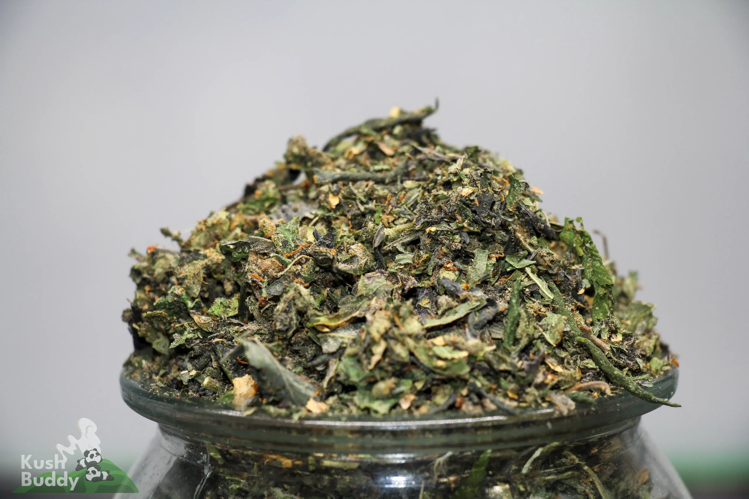 Close-up of a cannabis nugget piled on a glass jar, showing green leaves and frosty trichomes with orange hairs.