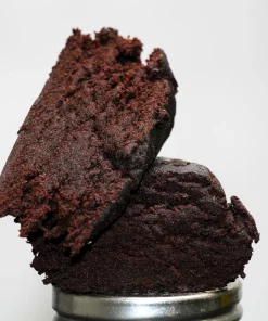 Close-up of dark, crumbly brownie pieces stacked on a metal lid against a light gray background.