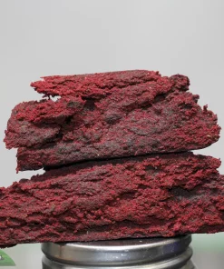 Stack of red cannabis concentrate pieces piled on a metal grinder, with a blurred background and a visible watermark in the corner.