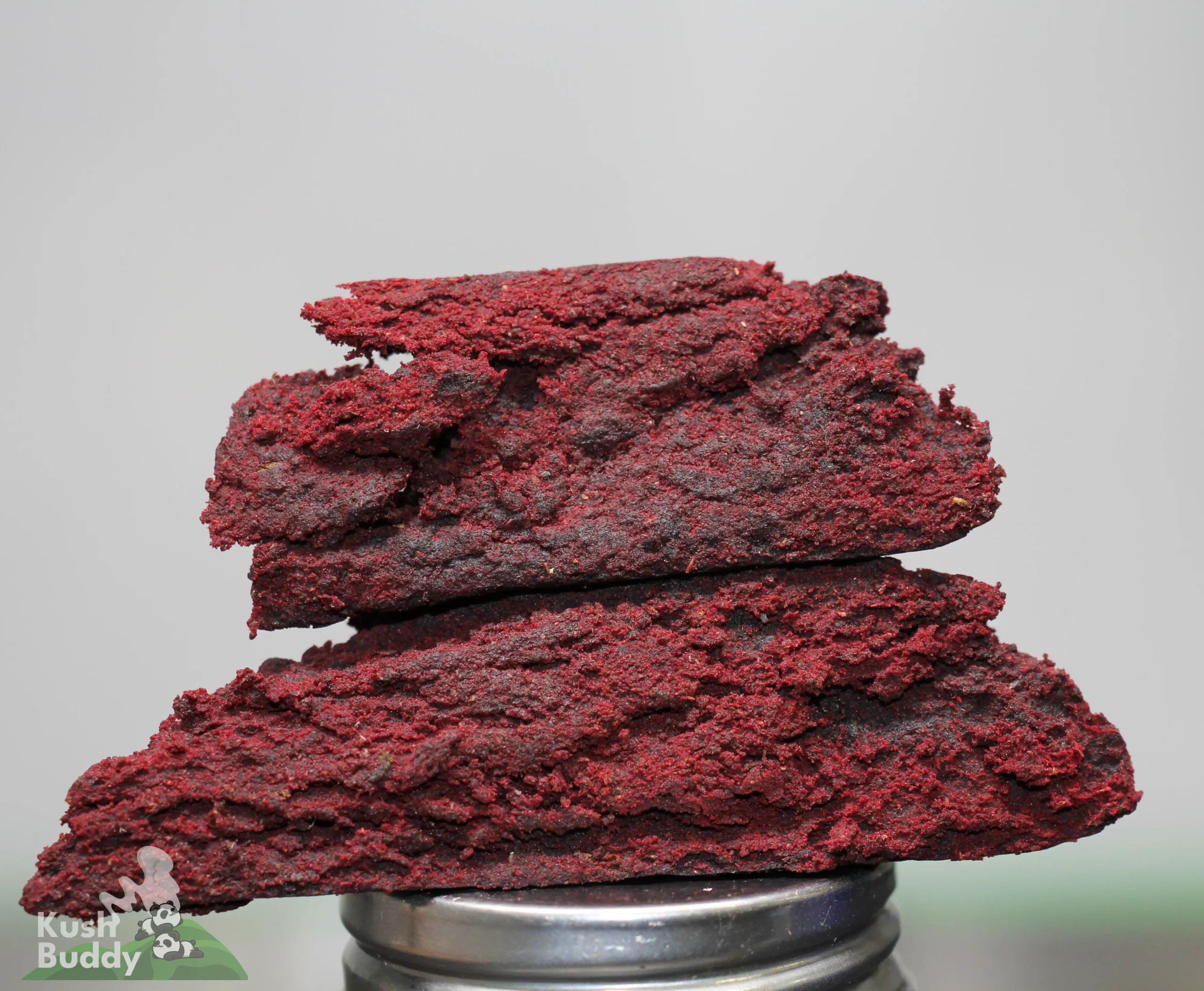 Stack of red cannabis concentrate pieces piled on a metal grinder, with a blurred background and a visible watermark in the corner.