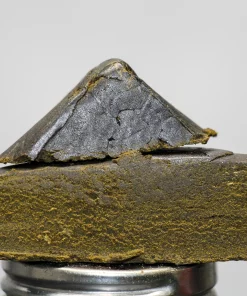 Close-up of a dark, metallic-looking rock slab stacked on a metal jar, with the Kush Buddy logo in the bottom-left corner.