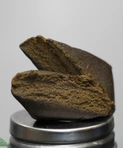 Close-up of dark brown, crumbly rocks stacked on a metal grinder, with Kush Buddy logo visible in the bottom-left corner.