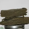 Stack of dark, crumbly cannabis bud slabs stacked on a metal grinder with the Kush Buddy watermark in the bottom-left corner.