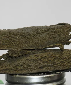 Stack of dark, crumbly cannabis bud slabs stacked on a metal grinder with the Kush Buddy watermark in the bottom-left corner.