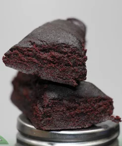 Close-up of a dark, crumbly chocolate brownie slice stacked on a metal grinder, with a faint logo in the corner.