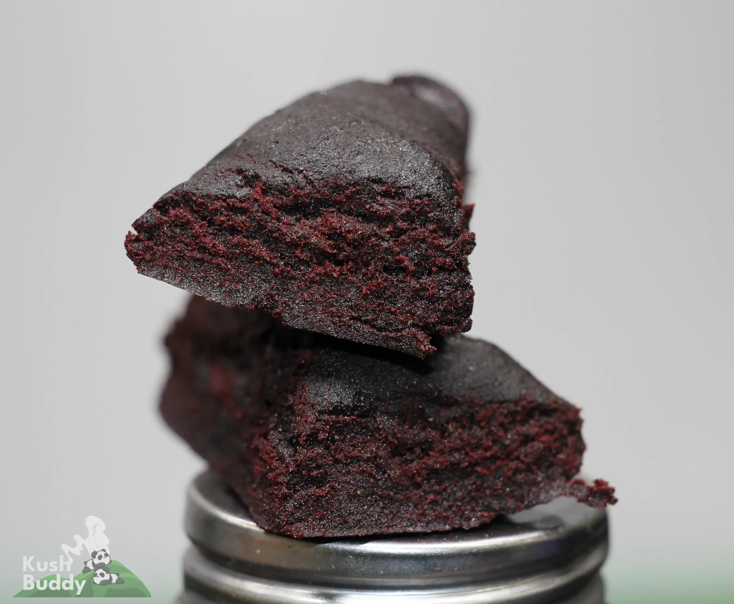 Close-up of a dark, crumbly chocolate brownie slice stacked on a metal grinder, with a faint logo in the corner.
