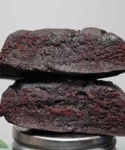 Close-up of dark red, crumbly cookies stacked on a metal lid against a pale gray background; watermark visible in the bottom-left corner.