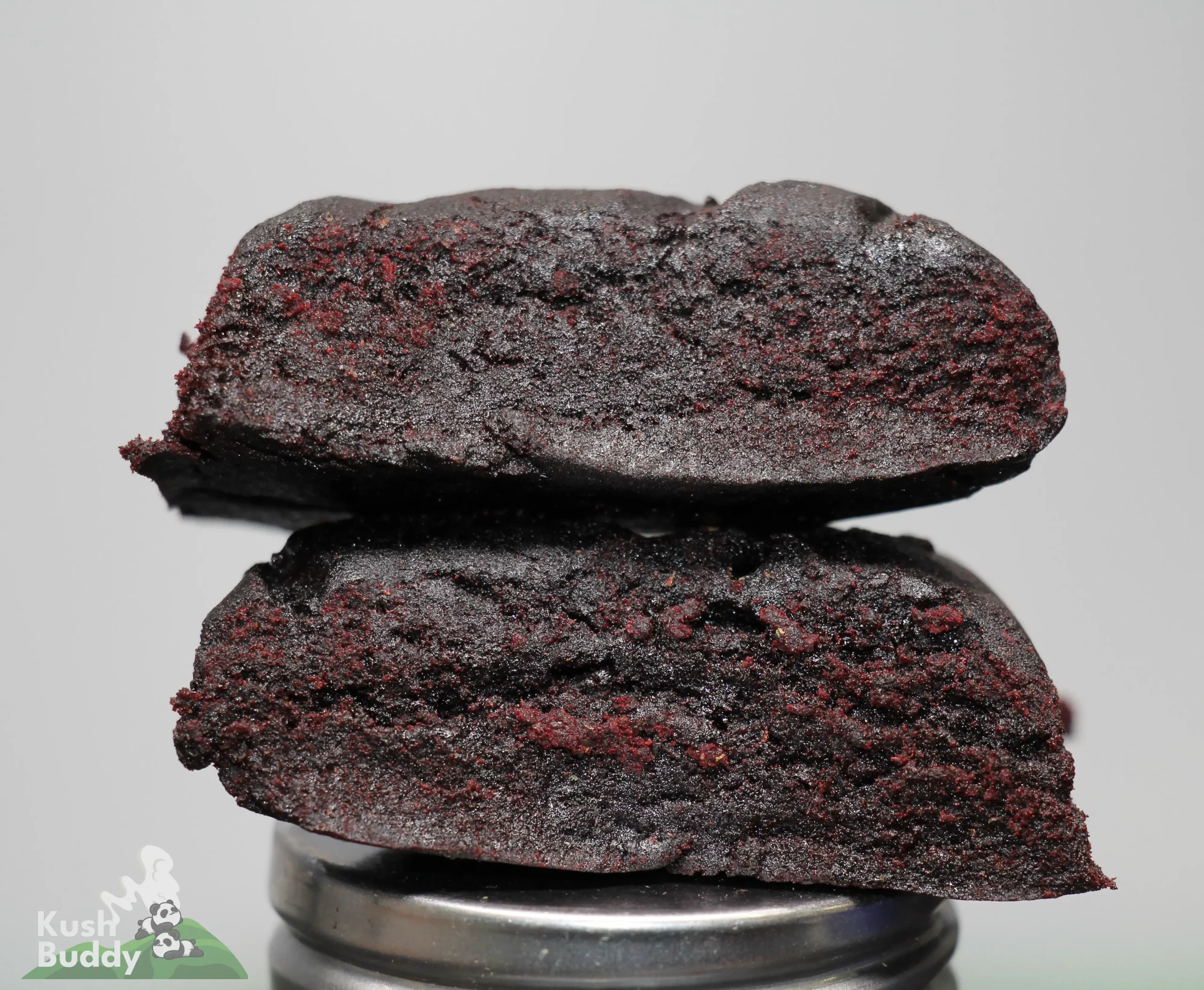 Close-up of dark red, crumbly cookies stacked on a metal lid against a pale gray background; watermark visible in the bottom-left corner.
