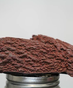 Close-up of dark red cannabis concentrate crumble resting on a metal grinder, with a Kush Buddy watermark in the corner.
