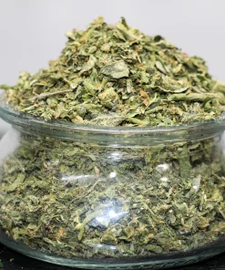 Pile of chopped cannabis buds in a clear glass jar, Kush Buddy watermark visible at the bottom left