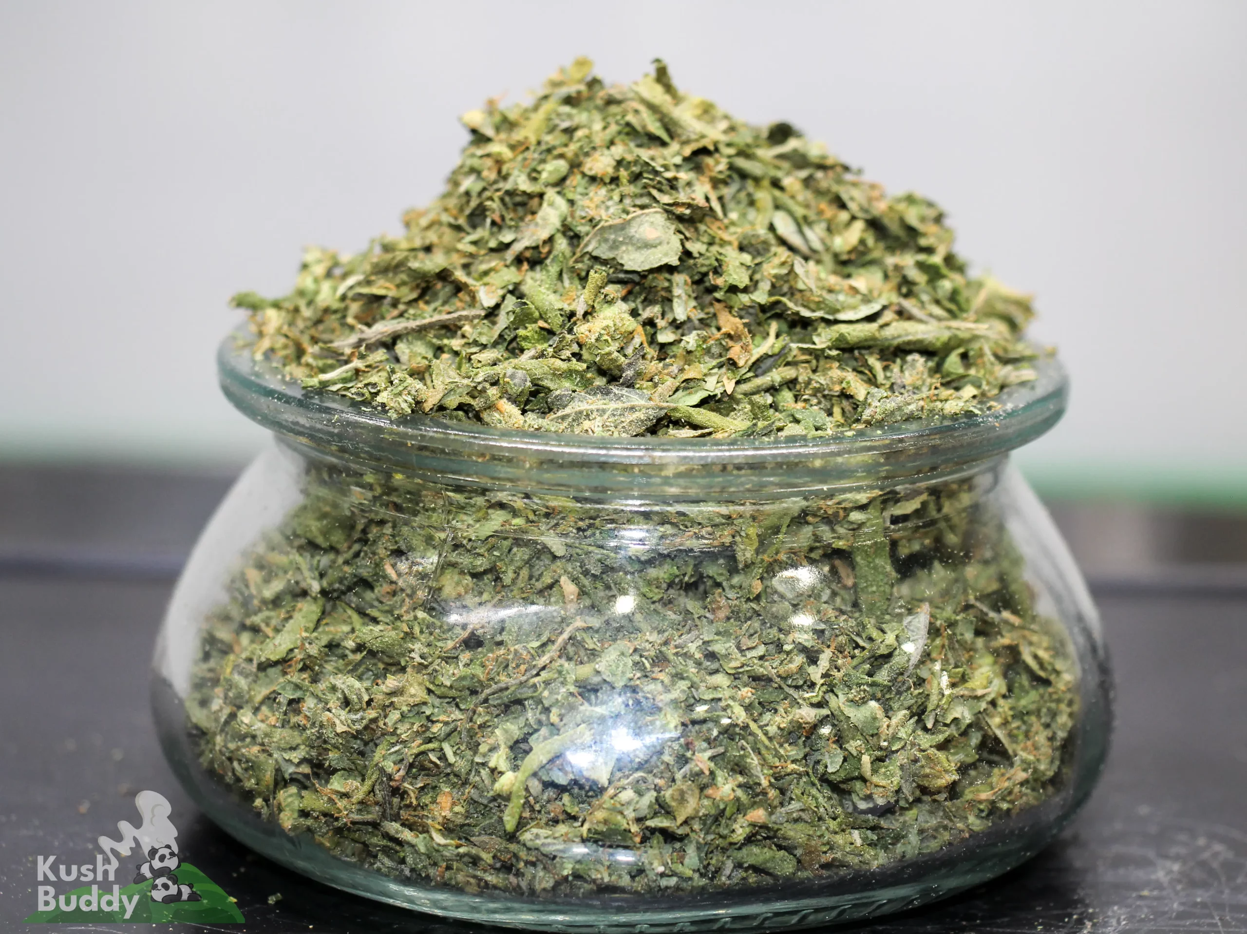 Pile of chopped cannabis buds in a clear glass jar, Kush Buddy watermark visible at the bottom left