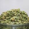 Close-up of a heap of chopped cannabis buds piled on a glass container, with a Kush Buddy watermark in the corner.