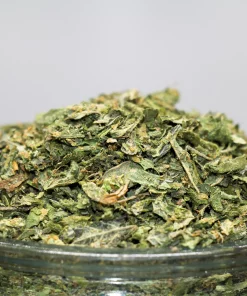 Close-up of a heap of chopped cannabis buds piled on a glass container, with a Kush Buddy watermark in the corner.