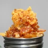 Close-up of amber cannabis concentrate crumble with crystal-like sugars stacked on a metal grinder, shown against a neutral background.