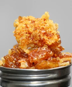 Close-up of amber cannabis concentrate crumble with crystal-like sugars stacked on a metal grinder, shown against a neutral background.