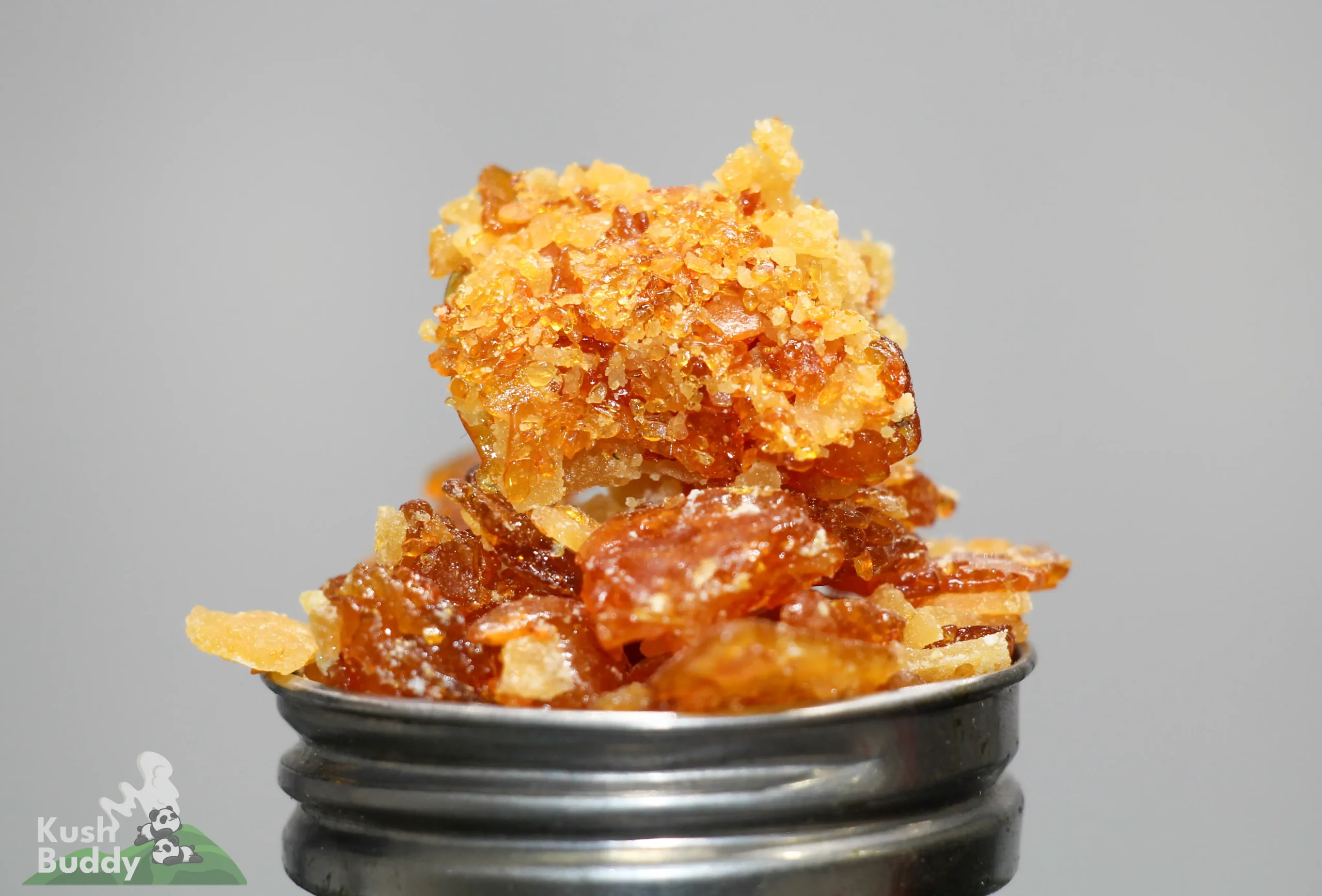 Close-up of amber cannabis concentrate crumble with crystal-like sugars stacked on a metal grinder, shown against a neutral background.
