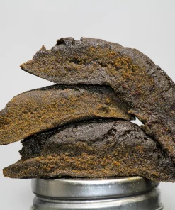 Stack of dark, crumbly cookies piled on a metal cap, with the Kush Buddy logo in the bottom-left corner.