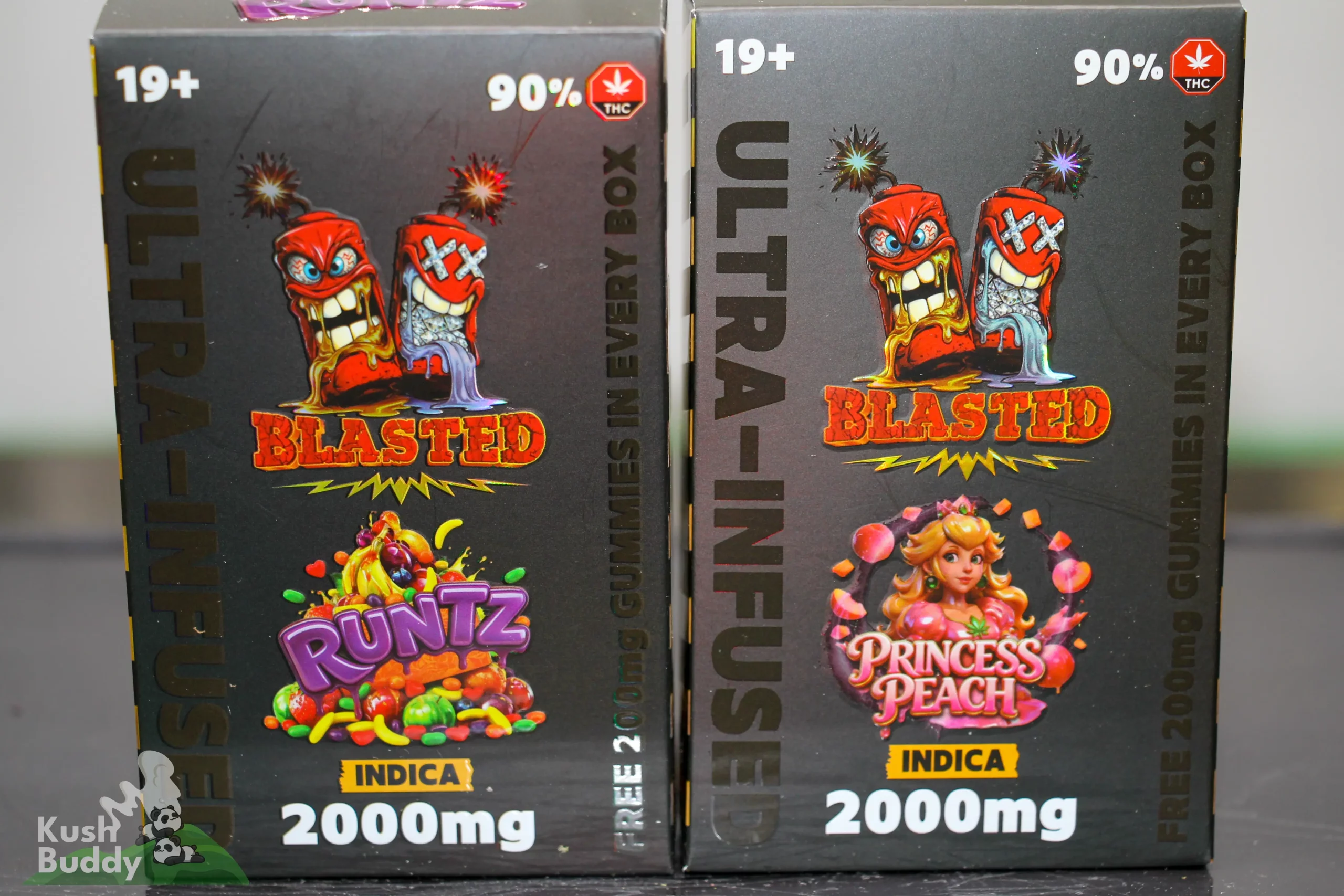 Two dark packaging boxes for cannabis products standing side by side, featuring cartoon red characters and the word BLASTED with INDICA and 2000mg text visible.