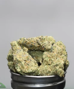 Close-up of cannabis buds stacked in a black container, with frosty trichomes and orange hairs on a gray background, watermark in corner.