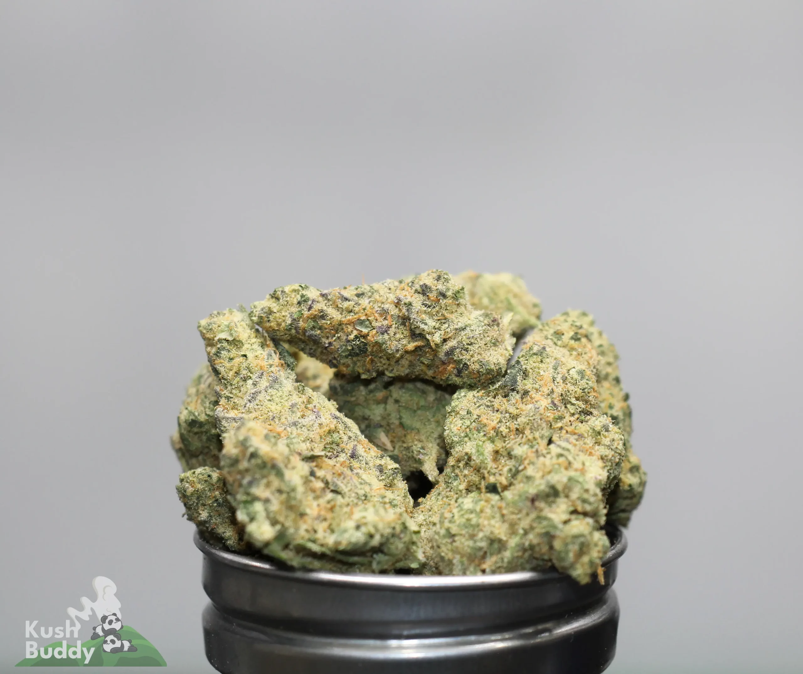 Close-up of cannabis buds stacked in a black container, with frosty trichomes and orange hairs on a gray background, watermark in corner.