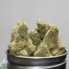 Close-up of frosty cannabis buds in a metal container with a Kush Buddy watermark in the corner.